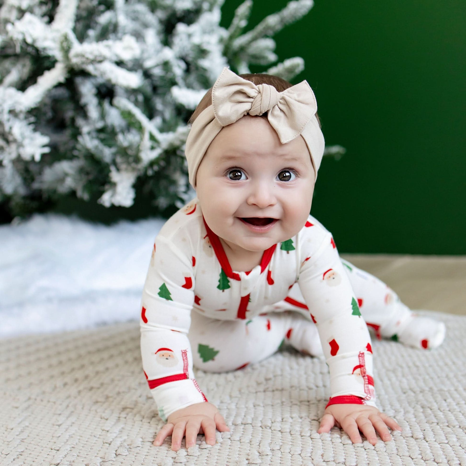 Baby in a Christmas-themed outfit with a bow headband, sitting on a textured surface with a snowy tree in the background.